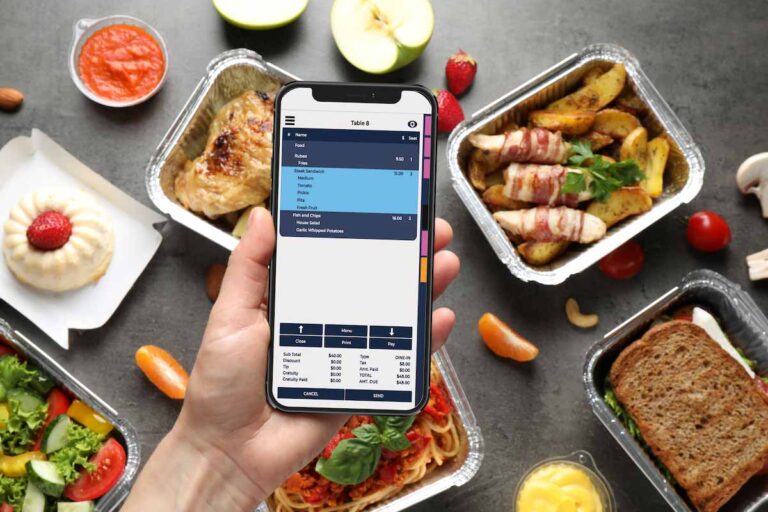 A person's hand is holding a smartphone displaying a mobile point-of-sale (POS) system interface, seemingly in a restaurant setting. The screen shows a customer's order with items like "Ruben," "Fries," and "Fish and Chips," along with their prices. Below the order, there's a breakdown of the subtotal, tax, tip, and grand total. Buttons for "Close," "Menu," "Amount," and payment options are also visible. Surrounding the hand and phone on a gray textured surface are several aluminum takeout containers filled with various food items: a piece of roasted chicken, bacon-wrapped potato wedges, a pasta dish with meat sauce, and a sandwich. There are also small dishes containing a red sauce and a yellow sauce, as well as fresh produce like sliced apples, strawberries, and cherry tomatoes scattered around, suggesting a variety of menu offerings. The scene implies the finalization of a takeout order using a mobile POS system.