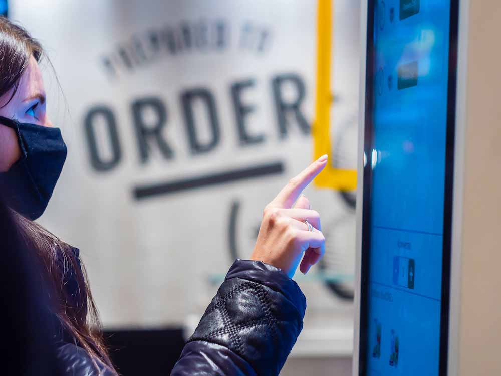 A person with long dark hair, wearing a black face mask and a black jacket with a quilted pattern on the sleeves, is interacting with a touch screen monitor. The screen has a light blue interface and displays the word "ORDER" in large letters. The person's right index finger is extended, seemingly touching the screen.