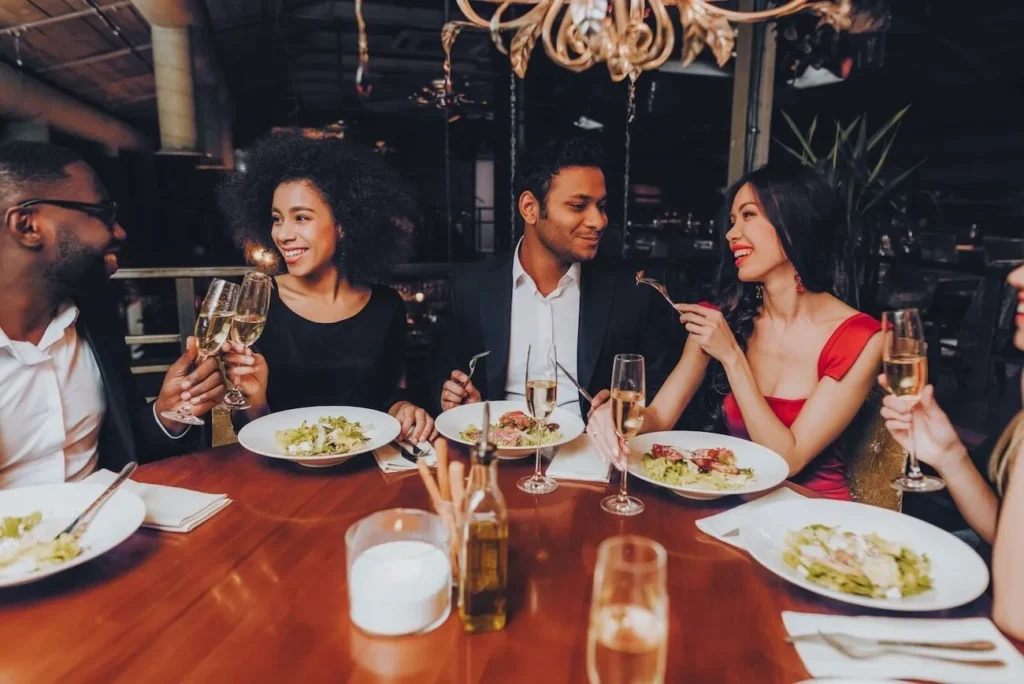 Five individuals are seated around a wooden table in a dimly lit, upscale restaurant. They appear to be enjoying a meal together, as evidenced by the plates of food, wine glasses, and cutlery in front of them. The person on the far left has dark skin and is wearing glasses and a light-colored shirt under a dark jacket. Next to them is a person with curly dark hair, smiling and holding a wine glass. Across the table sits a person with short dark hair, wearing a dark suit jacket and looking towards the person with curly hair. To their right is a person with long dark hair, wearing a red dress and holding a fork to their mouth while looking at the person in the suit. Part of another person is visible on the far right, holding a wine glass. The atmosphere suggests a social gathering or celebration.