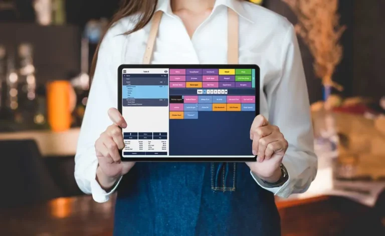 A person wearing a white long-sleeved shirt and a denim apron is holding a black tablet displaying a point-of-sale (POS) system interface. The screen shows various sections with different colors and text, likely representing menu items or functions. The person's hands are holding the tablet from the sides, with their thumbs visible on the screen's bezel. The background is blurred, suggesting a restaurant or cafe setting with warm lighting and indistinct shapes of tables and chairs. The focus is on the tablet and the hands holding it, emphasizing the use of technology in order taking or payment processing.